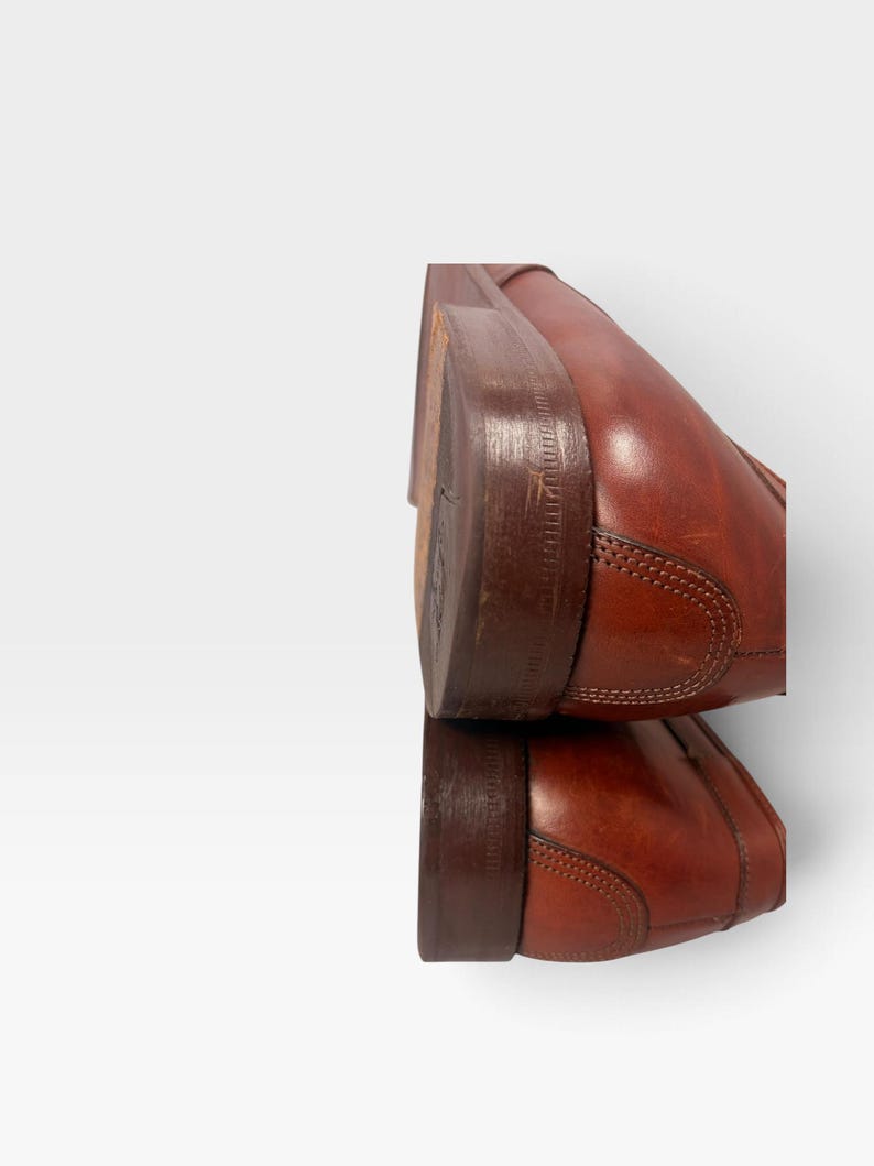 May include: A pair of stacked brown leather shoes featuring a smooth finish, visible stitching along the heel counter, and a dark brown stacked wooden heel. The shoes are positioned one above the other, showcasing the side profile and heel construction.