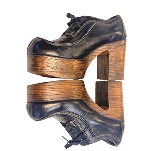 May include: A black lace-up shoe with a thick, chunky wooden platform sole and a high block heel. The upper part of the shoe is made of smooth black material with visible stitching details, while the base features a prominent wood grain texture.
