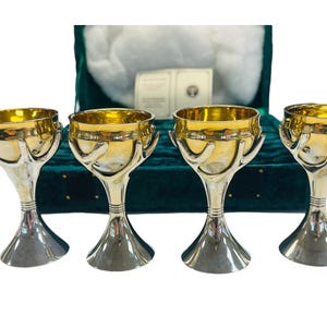 May include: Four silver and gold goblets with a unique design, featuring a gold interior and a silver exterior. The goblets are displayed in a green velvet-lined box. The goblets have a height of approximately 6 inches.