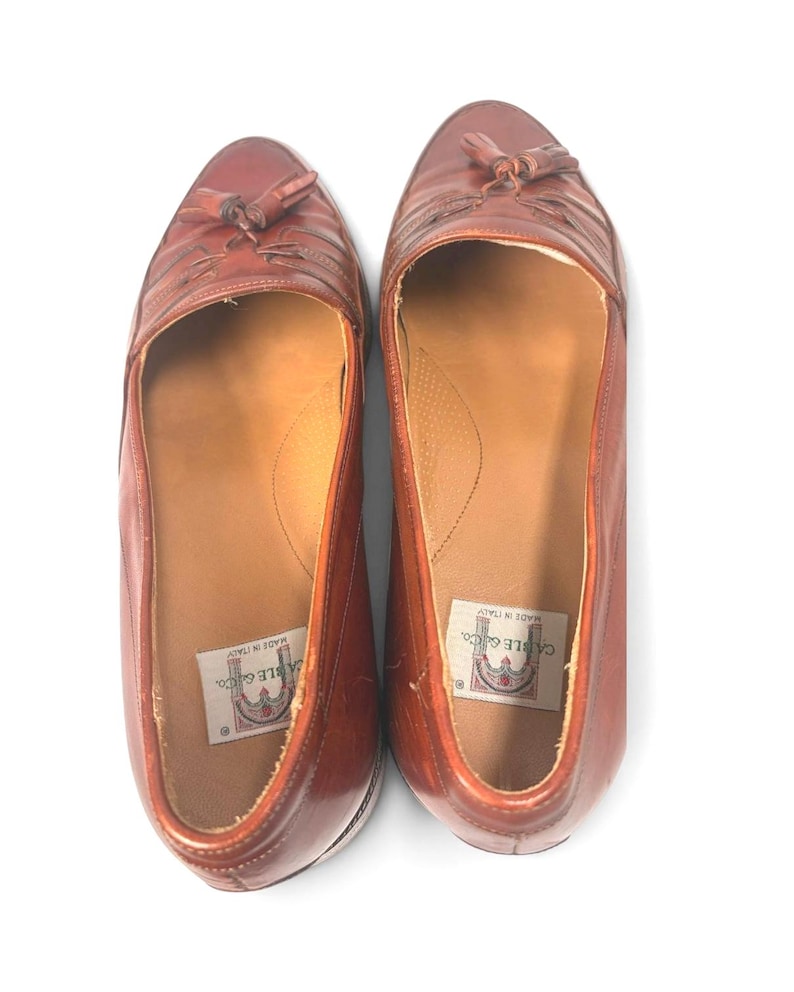 May include: A pair of brown leather loafers featuring a tassel detail on the top of each shoe. The interior of the shoes includes a white label with the text Cable & Co. and Made in Italy printed in black, visible inside the heel area.