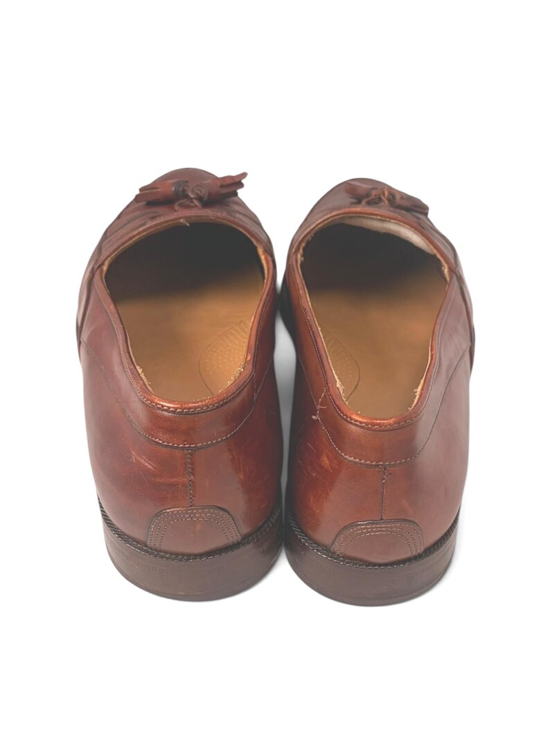 May include: A pair of brown leather loafers featuring a tassel detail on the top. The shoes have a smooth finish, a rounded toe, and a low heel. The interior is lined with a tan material, and the soles are constructed from a dark brown material.