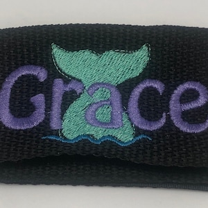 May include: Black fabric wristband with a purple embroidered name "Grace" and a green mermaid tail with a blue wave underneath.