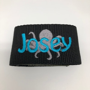 May include: Black fabric wristband with a gray octopus and the name "Josey" embroidered in blue.