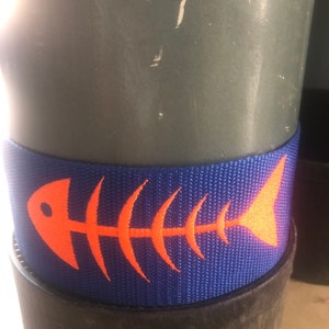May include: A blue and orange fish bone design on a blue nylon strap. The strap is wrapped around a gray cylinder.
