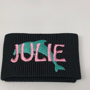 May include: Black fabric strap with pink embroidery that reads "JULIE" and a teal dolphin graphic.