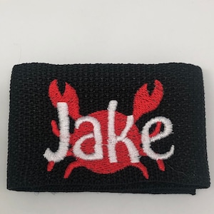 May include: Black fabric wristband with a red embroidered crab and the name "Jake" in white.