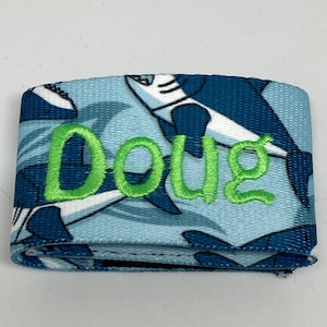 May include: A blue and white fabric wristband with a shark pattern and the name "Doug" embroidered in green.