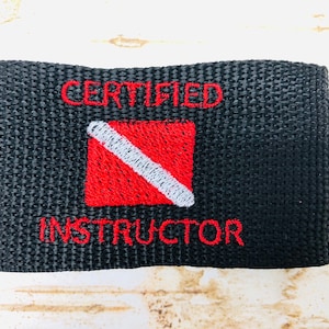 May include: Black rectangular patch with embroidered red text reading "CERTIFIED INSTRUCTOR." A red square with a white diagonal stripe is centered on the patch. The patch is likely for scuba diving certification.