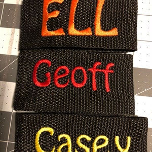 May include: Three black fabric wristbands with embroidered names in different colors. The top wristband has the name "ELL" embroidered in orange. The middle wristband has the name "Geoff" embroidered in red. The bottom wristband has the name "Casey" embroidered in yellow.