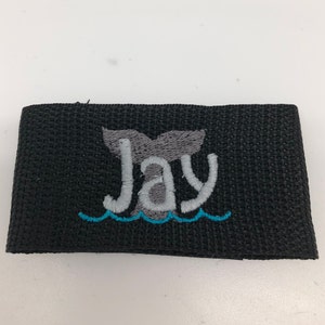May include: Black fabric wristband with a gray whale tail and the name "Jay" embroidered in white. The whale tail is above a blue wave.