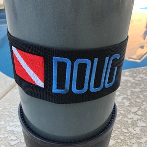 May include: A black scuba tank with a black and white scuba flag and the name "DOUG" embroidered in blue on a black strap.