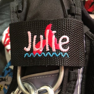 Puede incluir: Bolsa de tela negra con una etiqueta bordada en rosa y azul que dice "Julie". La etiqueta tiene un diseño de cola de sirena rosa.
