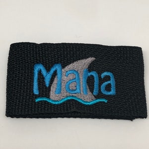 May include: Black fabric strap with a gray shark fin and blue waves embroidered on it. The word "Mana" is embroidered in blue and gray letters.