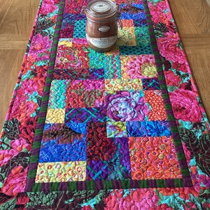 May include: A colorful patchwork table runner with a floral border. The runner is made of various fabrics in shades of pink, red, blue, green, and yellow. A glass jar with a lid is sitting on the runner. The jar has the text "Country Home" on it.