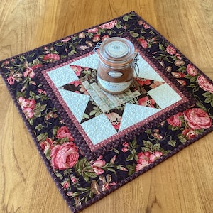 May include: A quilted table runner with a star pattern and floral print. The runner features a central star design in shades of brown, pink, and cream, surrounded by a border of dark purple fabric with pink rose patterns. A glass jar with a lid sits on top.