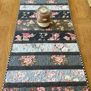 May include: A floral table runner with a patchwork design. The runner features alternating strips of patterned fabric in shades of grey, blue, and floral prints. A glass jar with a label sits on top. The edges have a dotted border.