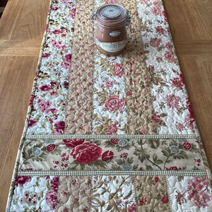 May include: A quilted table runner with a floral pattern in shades of pink, green, and beige. The runner has a gingham trim and is decorated with a glass jar labelled "Country Home".