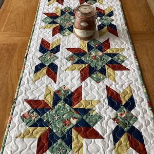 Peut inclure: Chemin de table matelassé avec des motifs d'étoiles rouges, marine, jaunes et verts sur fond blanc. Les centres des étoiles présentent des motifs floraux. Un pot avec le texte "COUNTRY HOME" est posé dessus.