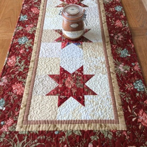 May include: A red and cream quilted table runner with a floral pattern and a star design. The runner has a brown gingham border and a jar of jam on top.