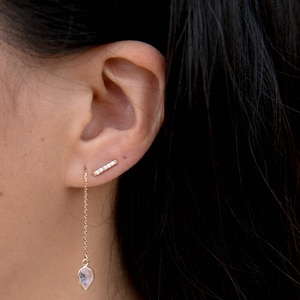 May include: A pair of gold earrings with a teardrop-shaped gemstone and a small bar of diamonds. The earrings are worn in a person's ear.