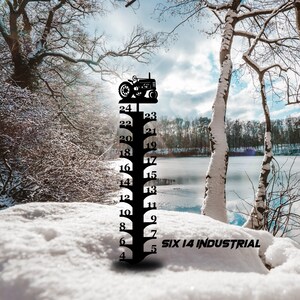 May include: Black metal snow gauge with a tractor silhouette at the top. The gauge measures up to 24 inches of snow. The text "SIX 14 INDUSTRIAL" is visible at the bottom. The gauge is set in a snowy landscape.