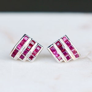 Puede incluir: Un par de pendientes de plata con un diseño geométrico que presenta múltiples filas de piedras de rubí de forma cuadrada.