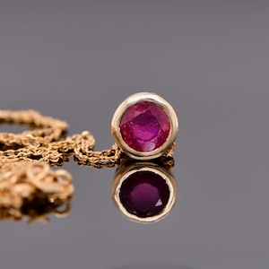 Puede incluir: Un collar de oro con un colgante de rubí redondo. El rubí es de color rosa intenso, engastado en un bisel. La cadena es de diseño delicado y enlazado. El colgante y la cadena están sobre una superficie reflectante.