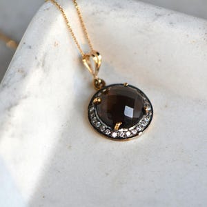 Könnte beinhalten: Eine goldene Halskette mit einem runden Anhänger, der einen großen, dunkelbraunen Edelstein zeigt, der von einem Heiligenschein aus kleineren, klaren Edelsteinen umgeben ist.