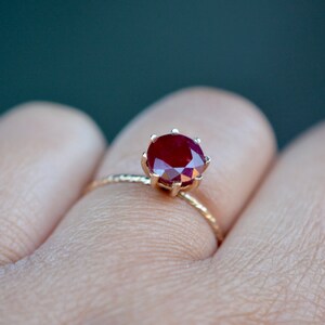 May include: A gold ring with a red ruby gemstone set in a prong setting. The ring is on a finger.