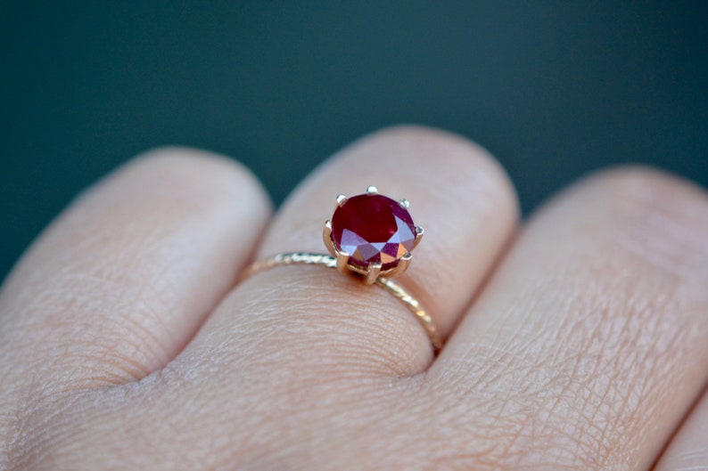 May include: A gold ring with a red ruby gemstone set in a prong setting. The ring is on a finger.
