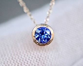 May include: A gold necklace featuring a round, faceted blue gemstone set in a bezel setting. The pendant is suspended from a delicate gold chain, showcasing the vibrant blue color and the reflective quality of the stone's cut and polished surface.