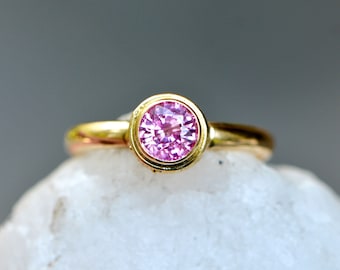 Puede incluir: Un anillo de oro con una piedra preciosa rosa redonda y facetada en un engaste de bisel circular liso. La banda es de oro pulido y la piedra central muestra un tono rosa vibrante con facetas visibles que captan la luz con brillo.