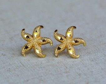 May include: A pair of gold-colored starfish stud earrings. Each earring features a five-pointed design with textured details and dark brown accents. The earrings are displayed on a neutral-colored fabric.