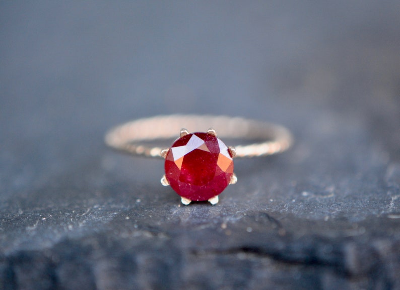 May include: A close-up of a ring with a red ruby gemstone set in a gold band.