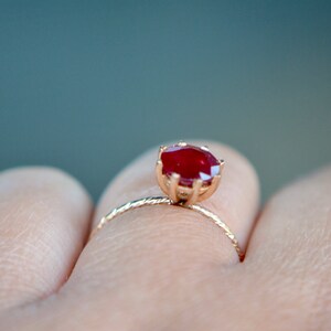 May include: A gold ring with a red gemstone set in a prong setting. The ring is on a finger.