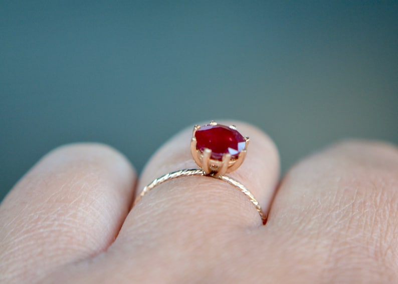 May include: A gold ring with a red gemstone set in a prong setting. The ring is on a finger.