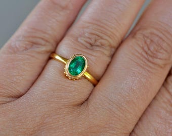 May include: A gold ring featuring an oval-cut, vibrant green gemstone. The ring's band is slender, and the gemstone is set in a detailed, gold bezel. The ring is displayed on a person's finger, showcasing its design and color.