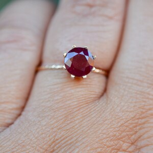 May include: A gold ring with a large, round, red ruby gemstone set in a prong setting.