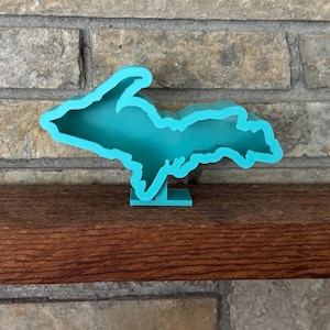 May include: A bright turquoise, three-dimensional plastic object shaped like the Upper Peninsula of Michigan. The item features a hollow interior and is mounted on a small, rectangular base, allowing it to stand upright on a flat surface.