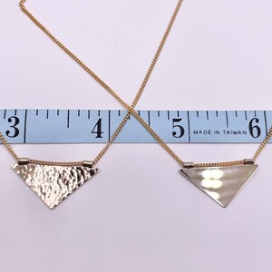 May include: Two gold necklaces with triangle pendants. The top necklace has a hammered triangle pendant and the bottom necklace has a smooth triangle pendant. Both necklaces are measured on a ruler that reads "Made in Taiwan" and shows inches.