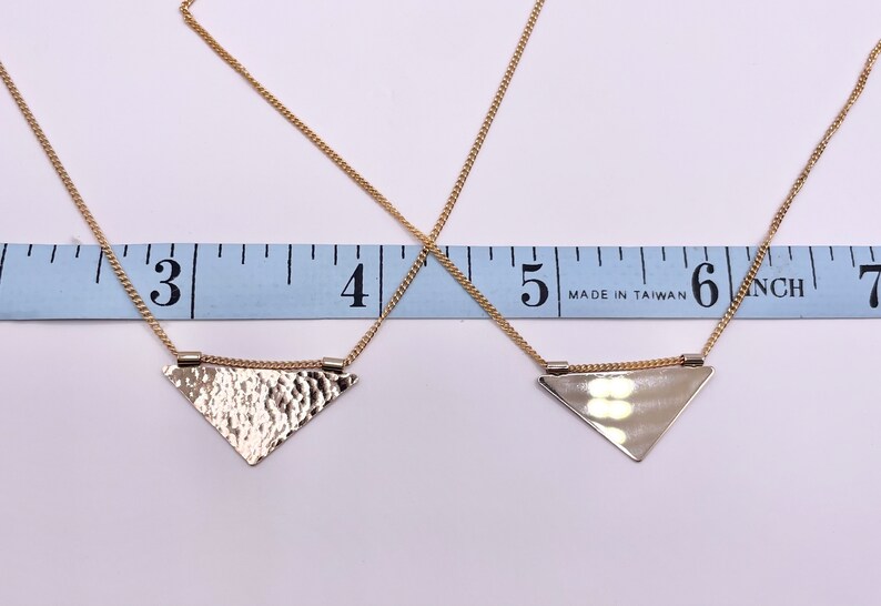 May include: Two gold necklaces with triangle pendants. The top necklace has a hammered triangle pendant and the bottom necklace has a smooth triangle pendant. Both necklaces are measured on a ruler that reads "Made in Taiwan" and shows inches.