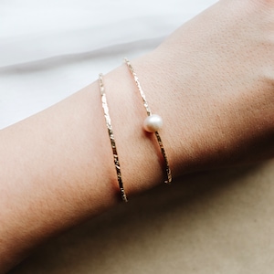 May include: Two gold bracelets with a textured finish, one with a single white pearl accent.