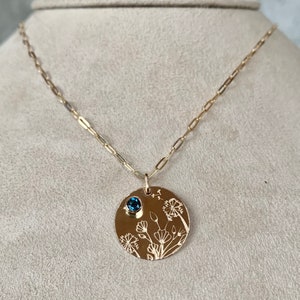 Peut inclure: Un collier doré avec un pendentif rond orné de motifs floraux gravés et d'une petite pierre précieuse bleue. Le pendentif est suspendu à une chaîne délicate. Le motif floral comprend des pissenlits et d'autres fleurs sauvages.