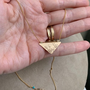 May include: A gold necklace with a triangular pendant featuring a delicate floral design. The pendant is attached to a thin gold chain with a turquoise bead accent.