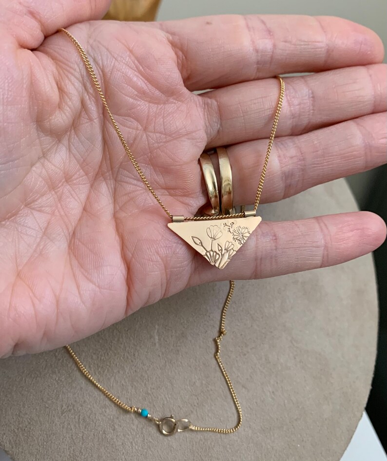 May include: A gold necklace with a triangular pendant featuring a delicate floral design. The pendant is attached to a thin gold chain with a turquoise bead accent.