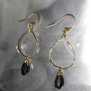 May include: Gold teardrop earrings with a dark brown gemstone pendant. The earrings are made of wire and have a delicate, hammered finish.