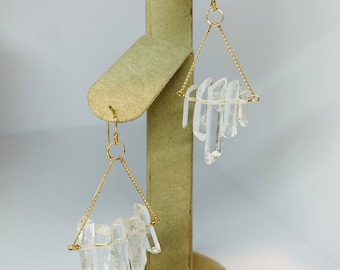 May include: Gold earrings with a delicate chain and clear quartz crystals hanging from the bottom. The crystals are arranged in a cluster and appear to be raw and unpolished.