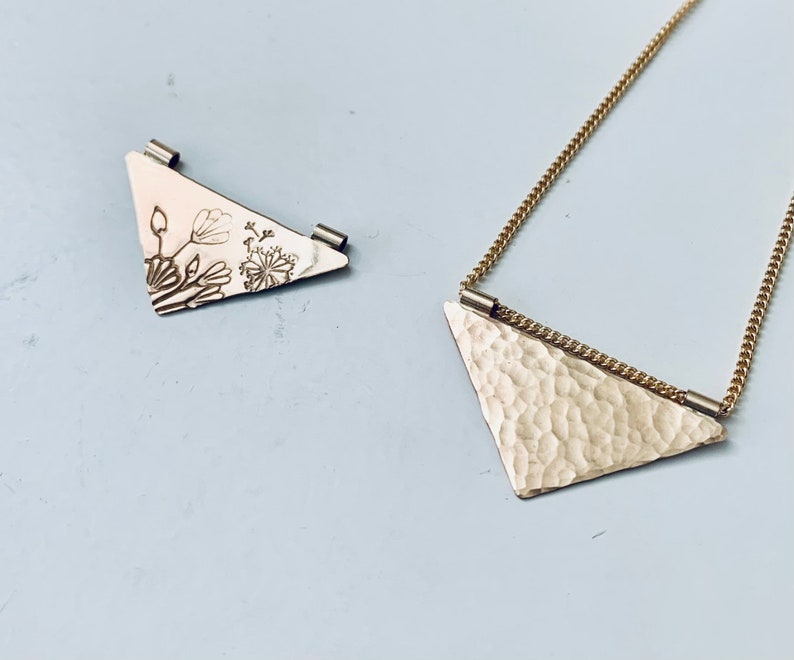 May include: Two gold triangle pendants, one with a floral design and the other with a hammered texture, on a gold chain.