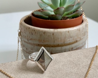Puede incluir: Un anillo de plata con una piedra cuadrada de color gris oscuro engastada en un engaste de plata. El anillo está sobre una superficie de ante marrón claro.