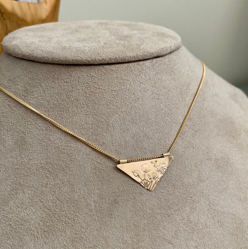 May include: A gold necklace with a triangular pendant featuring a delicate floral design. The pendant is attached to a thin gold chain.